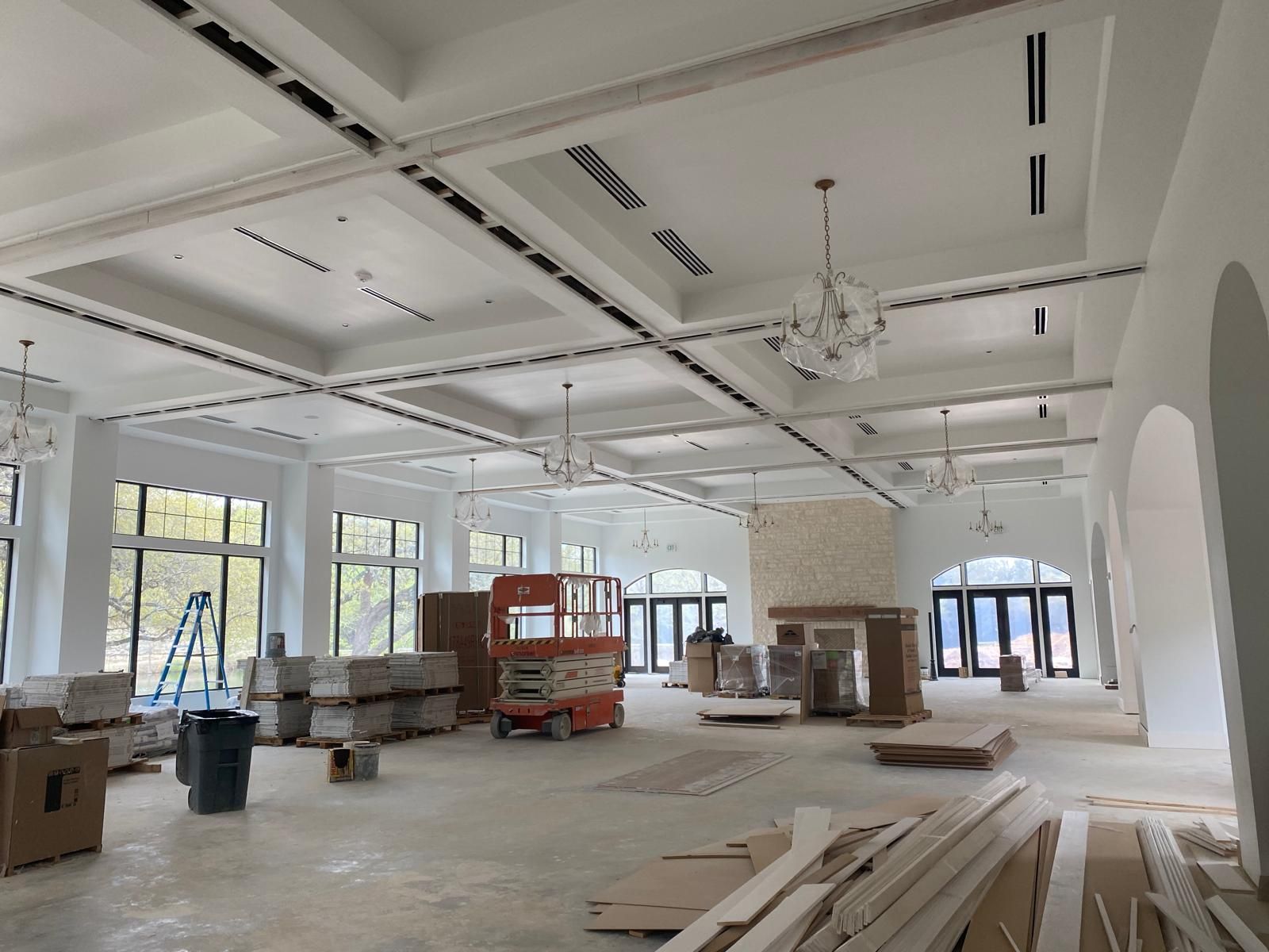 A large, bright, open-plan room under construction with vaulted ceilings, chandeliers, construction equipment, and boxes.