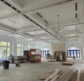 A large, unfinished interior space with white coffered ceilings, chandeliers, construction equipment, and stacked boxes.