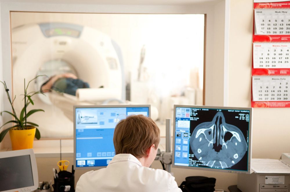 Un médico está mirando una pantalla de computadora mientras un paciente se somete a una tomografía computarizada.