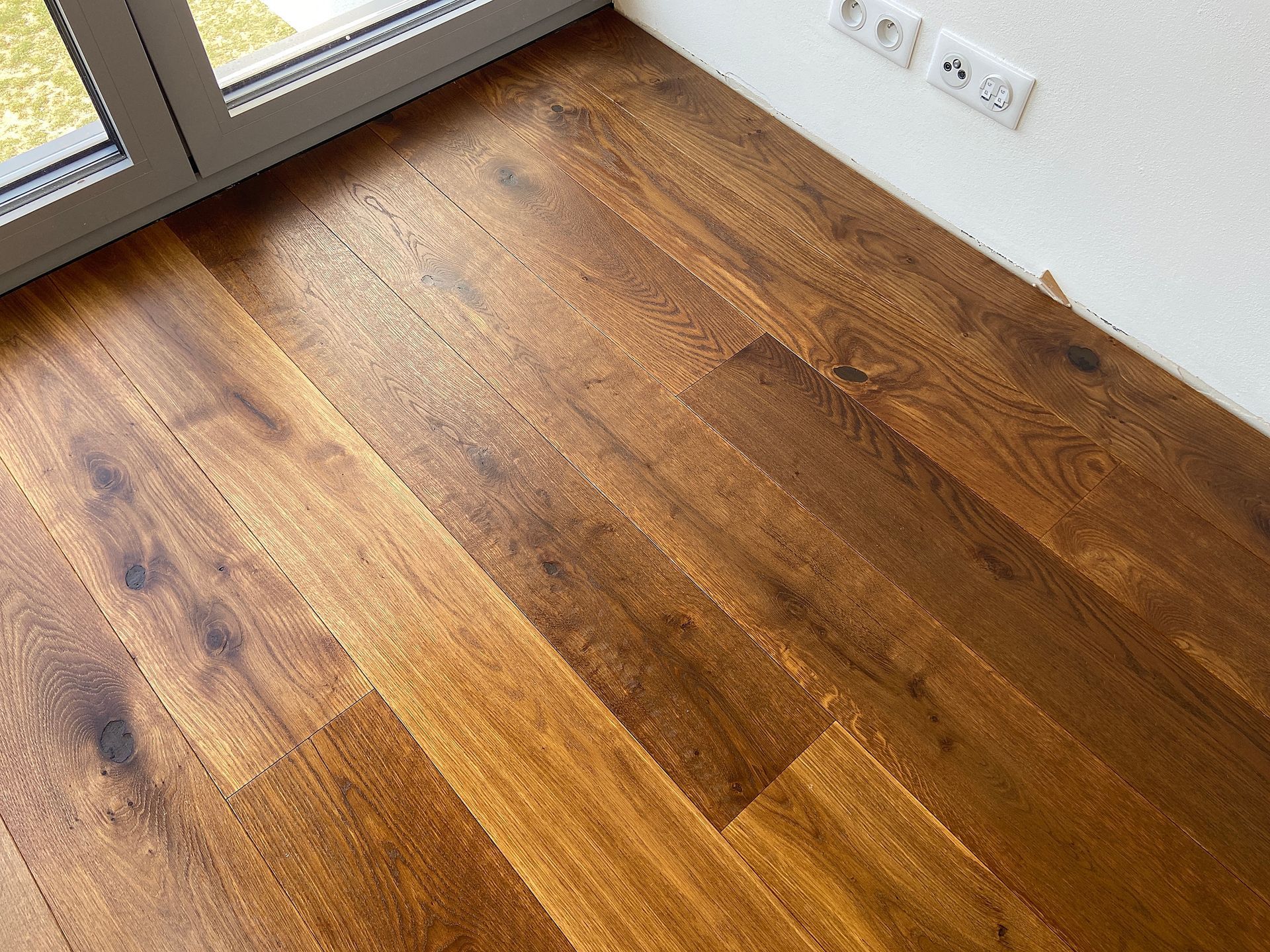 Wooden floor beside a glass door in a bright room with a white wall and electrical outlets.