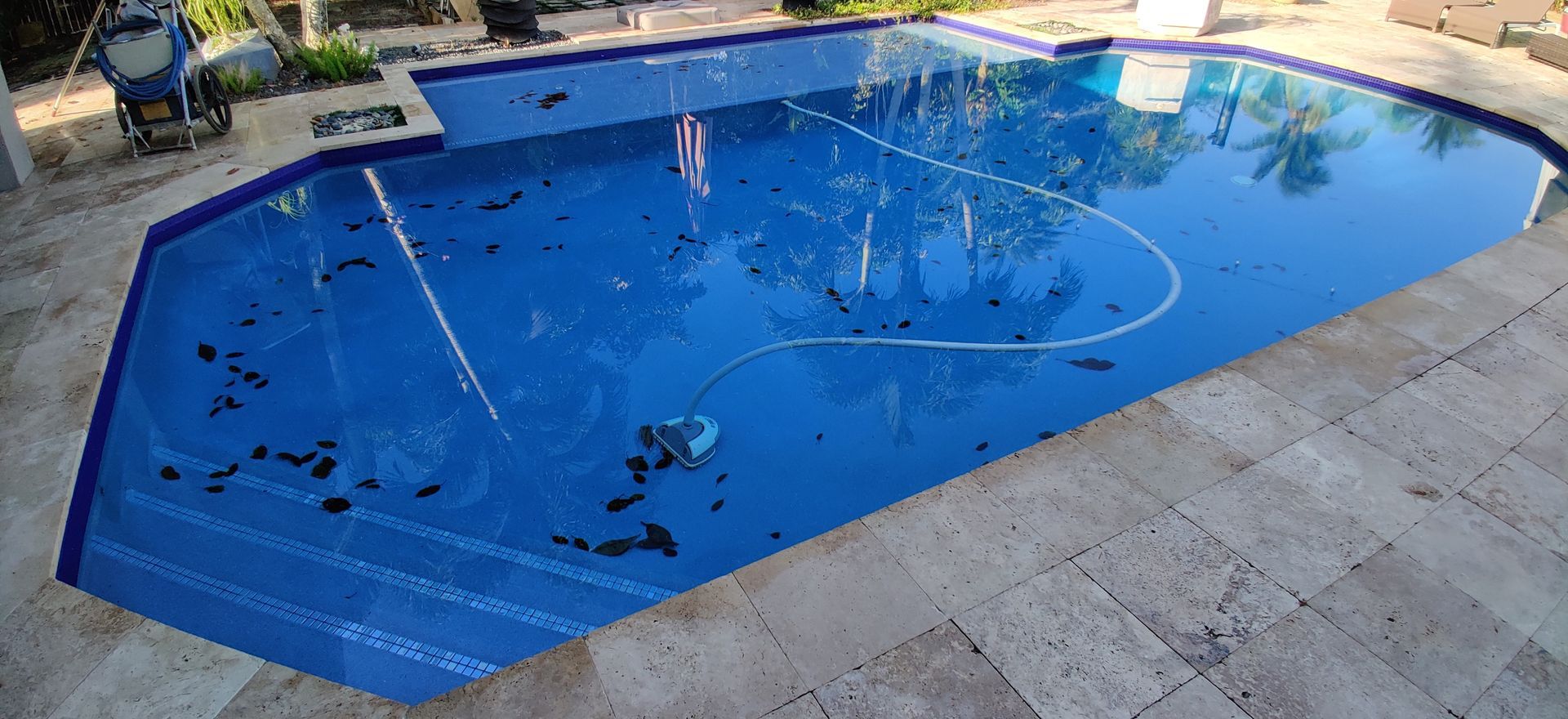 A blue swimming pool with a cleaner, surrounded by light-colored stone.