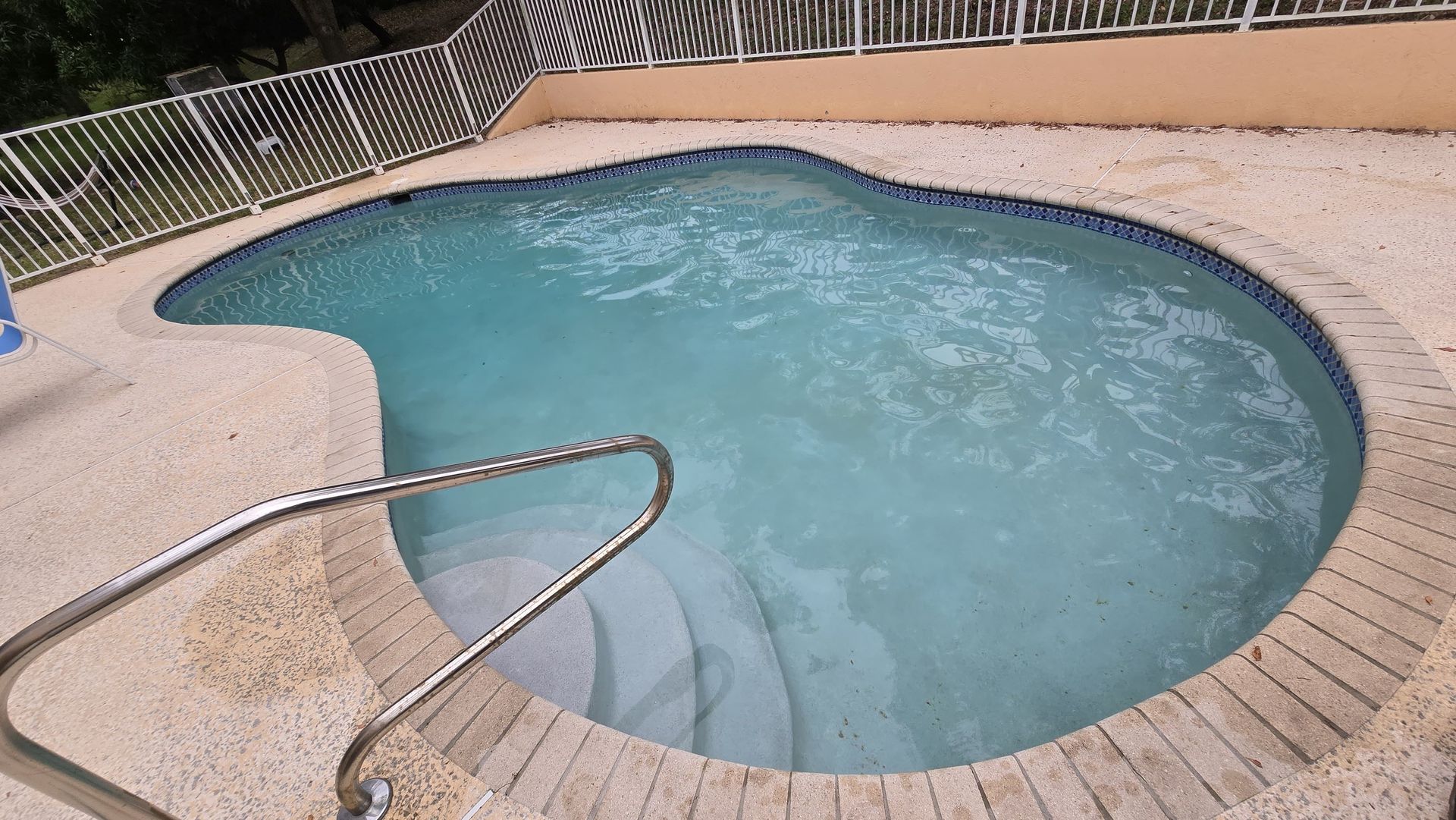 Small pool with blue water, steps, and a metal handrail. Tan brick surround, white fence in the background.