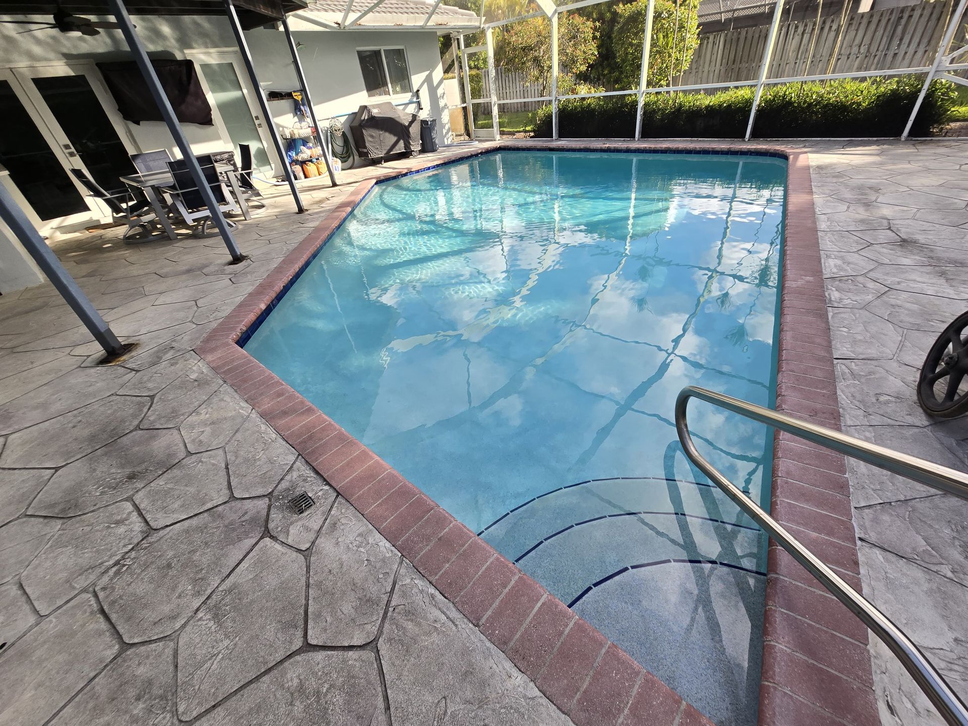 Swimming pool with blue water, brick border, and stone patio. Handrail leads into the pool.