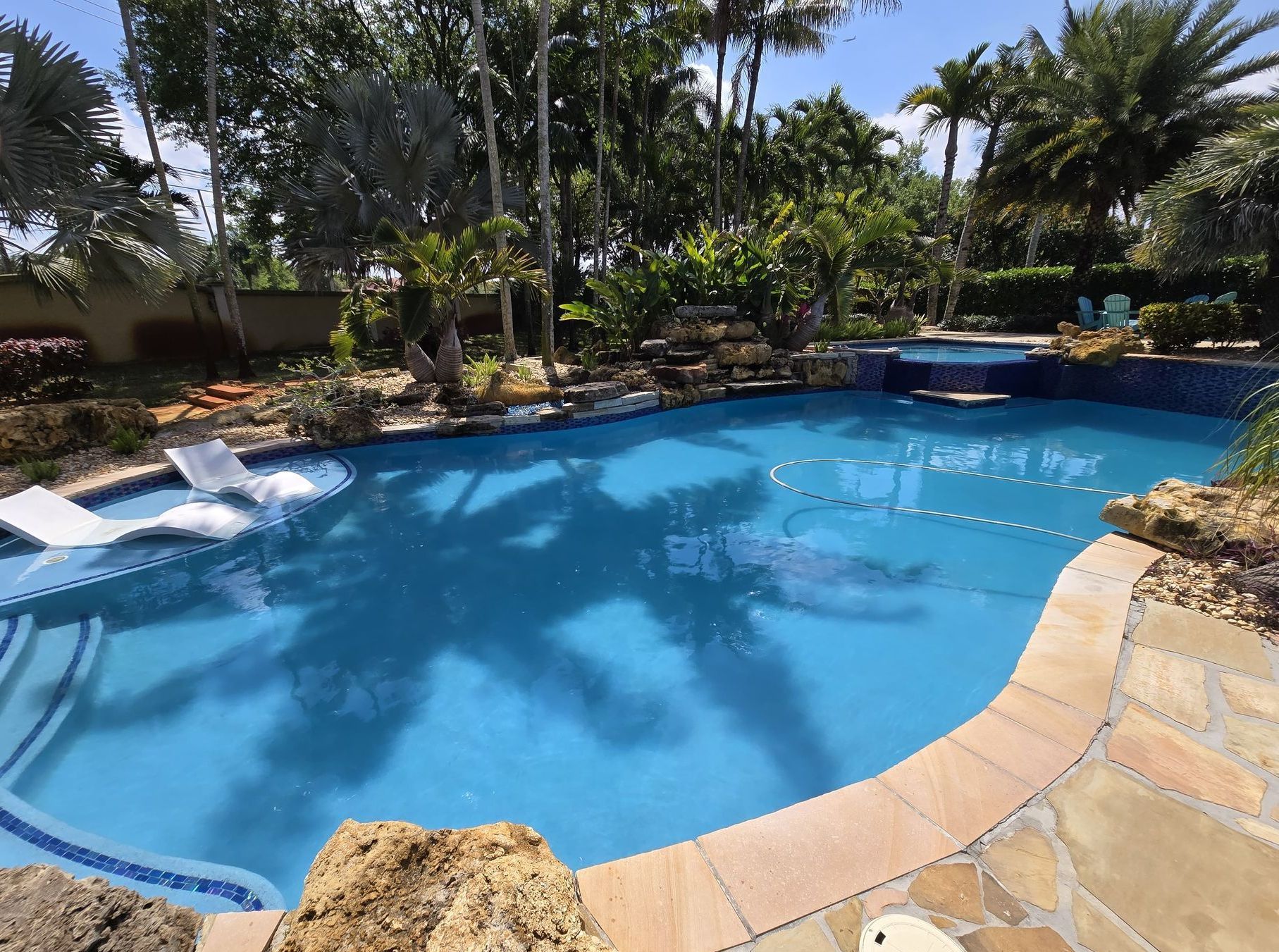 A backyard pool with a blue tiled surface, a waterfall, and lounge chairs. Palm tree shadows are cast on the water.