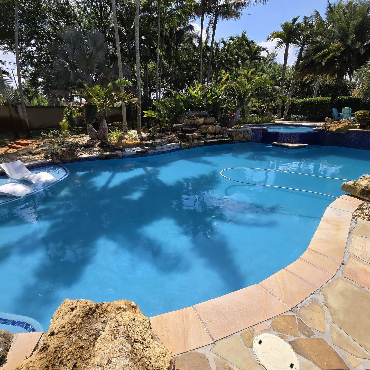 A blue swimming pool surrounded by stone and lush greenery on a sunny day.