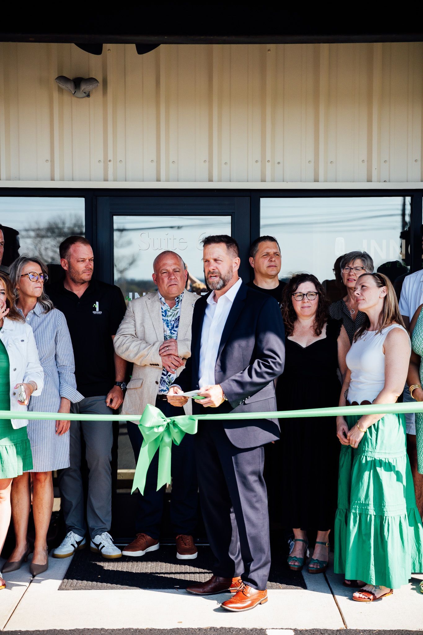 Ribbon-cutting at a building entrance, with a group of people standing behind a green ribbon and the speaker in front