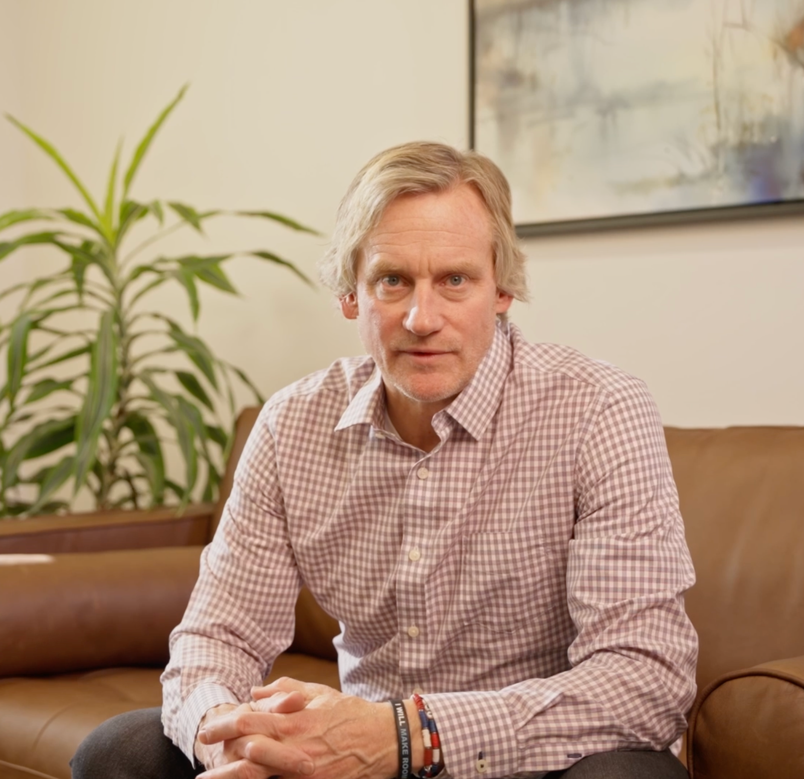 Man sitting on a brown couch in a bright living room, wearing a checkered shirt and looking at the camera.
