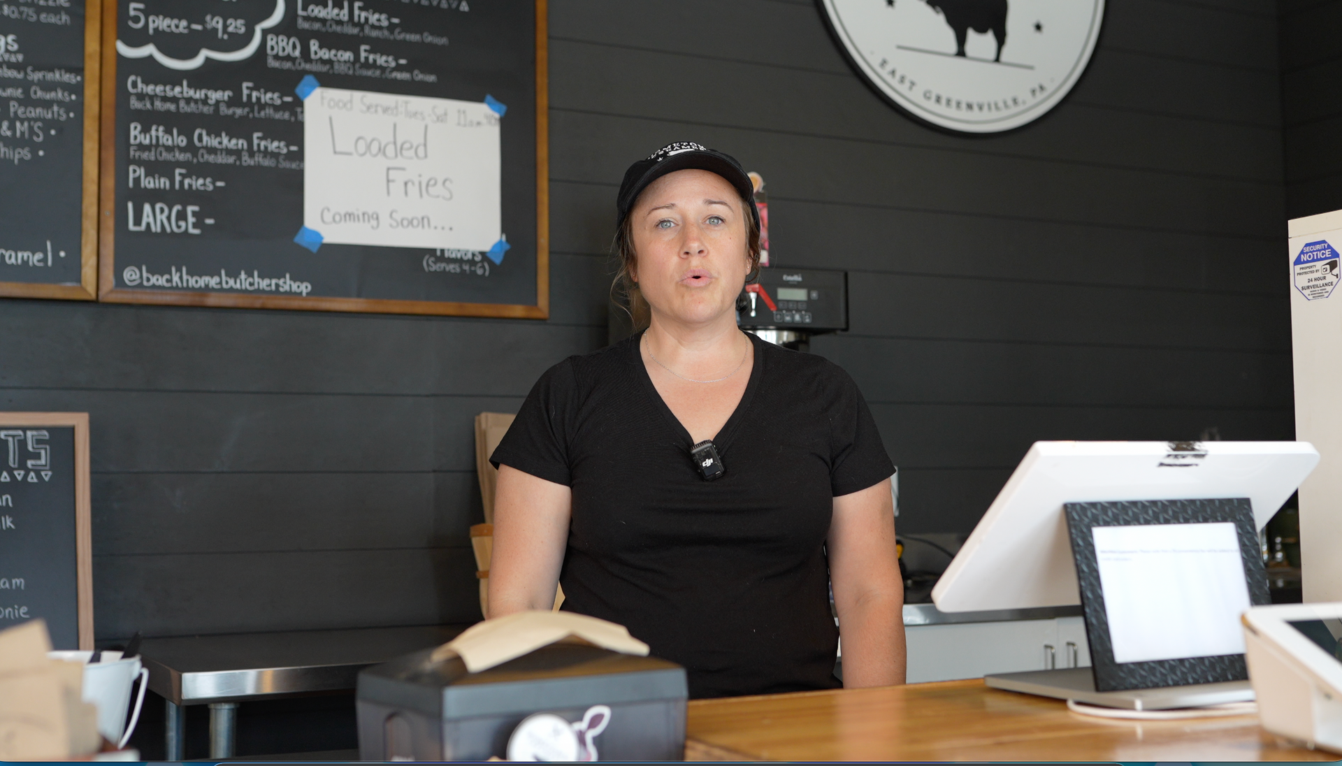 Woman behind a café counter with a register, white tablet stand, and chalkboard menu on a dark wall.