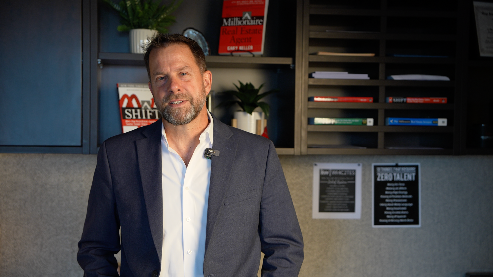 Man standing in a modern office, wearing a dark blazer over a white shirt, with shelves and framed signs behind him.
