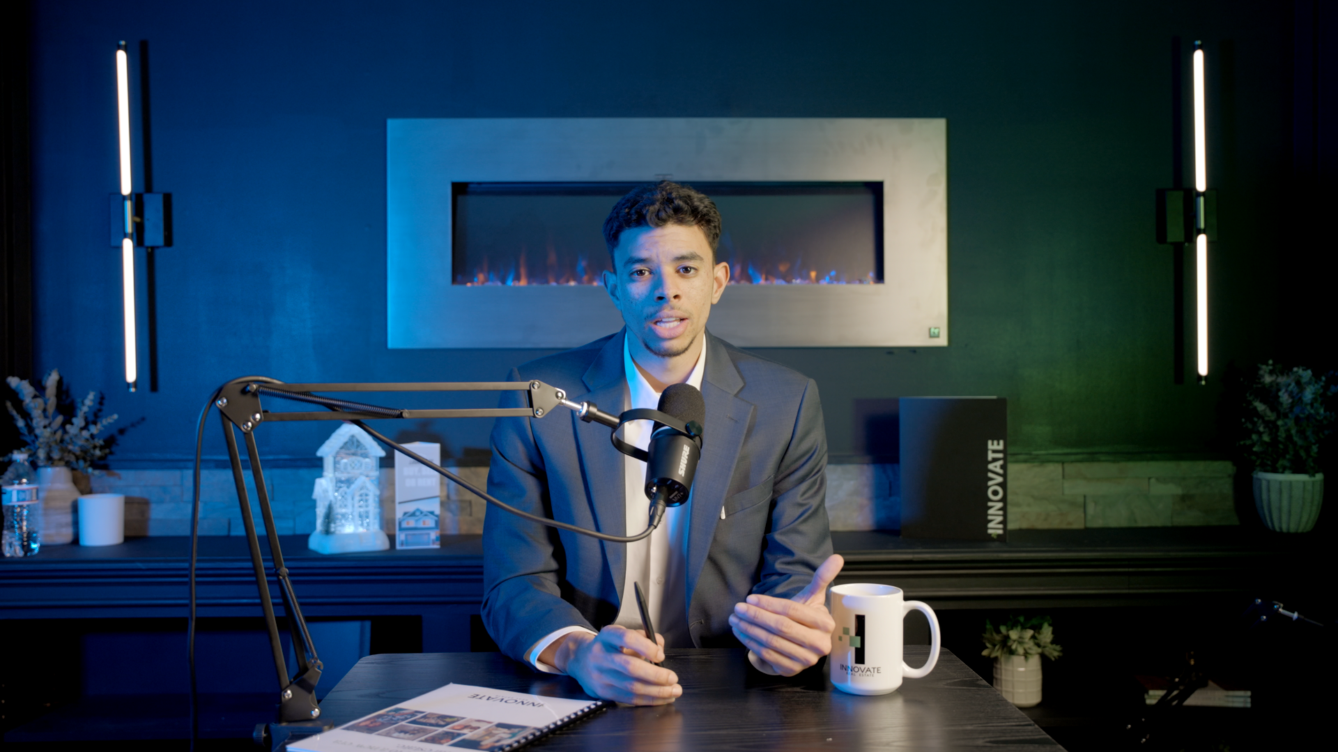 Person in a blue suit speaking into a microphone at a desk in a lit studio, with papers and a mug nearby.