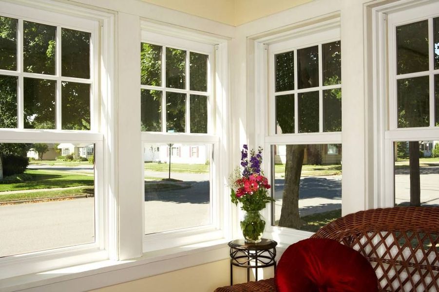 Corner room with white-framed windows overlooking a street. Flowers sit on a small table near a wicker chair.