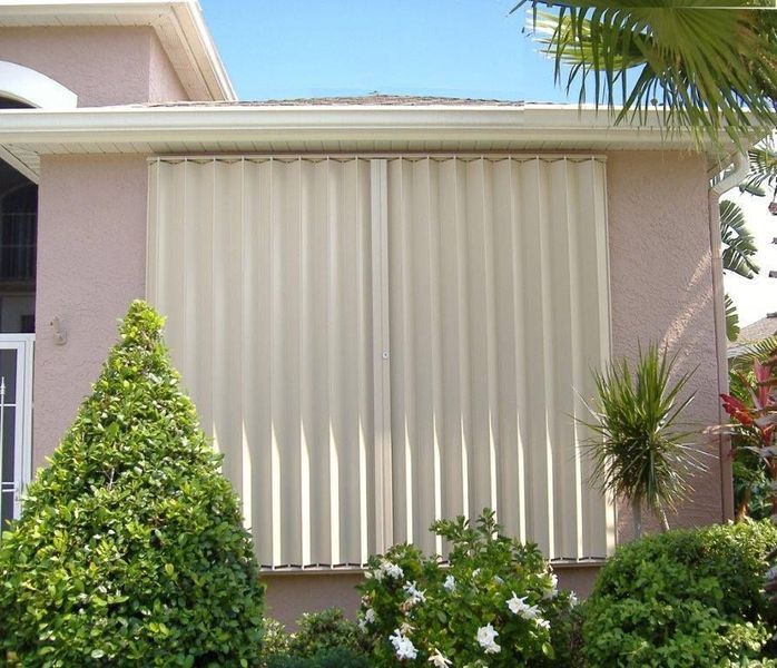Beige hurricane shutters protect a home from the elements, surrounded by greenery.