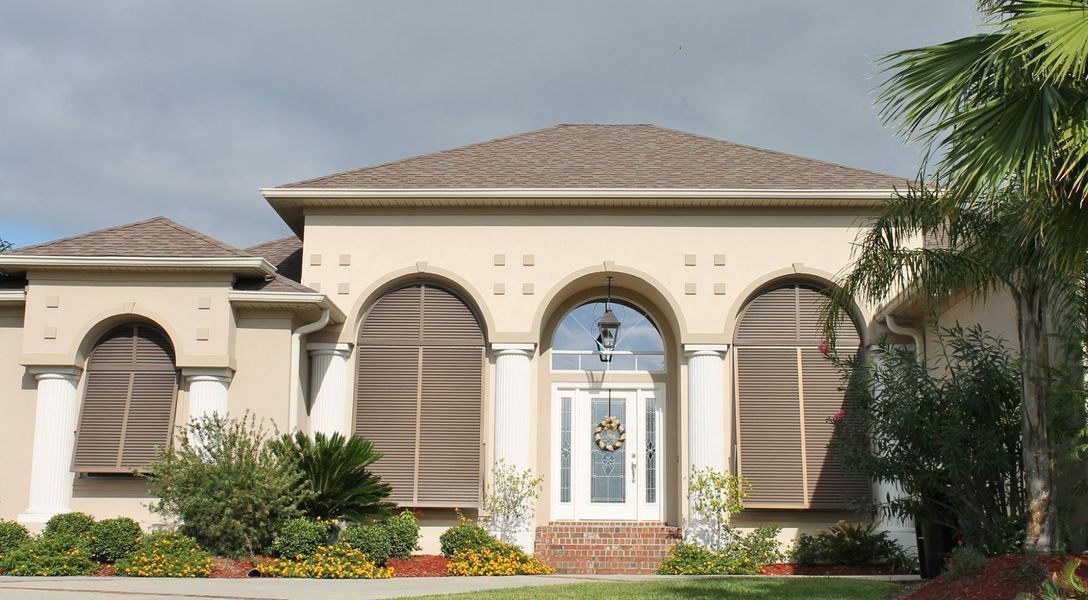 Beige stucco house with brown shutters and arched doorway; tropical plants in front.