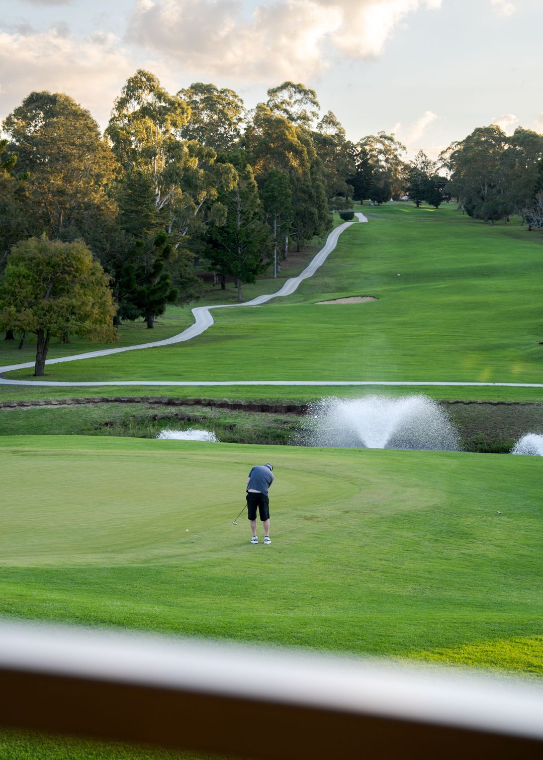 Golfer putting on a green near a water fountain, with a winding fairway and trees in the background