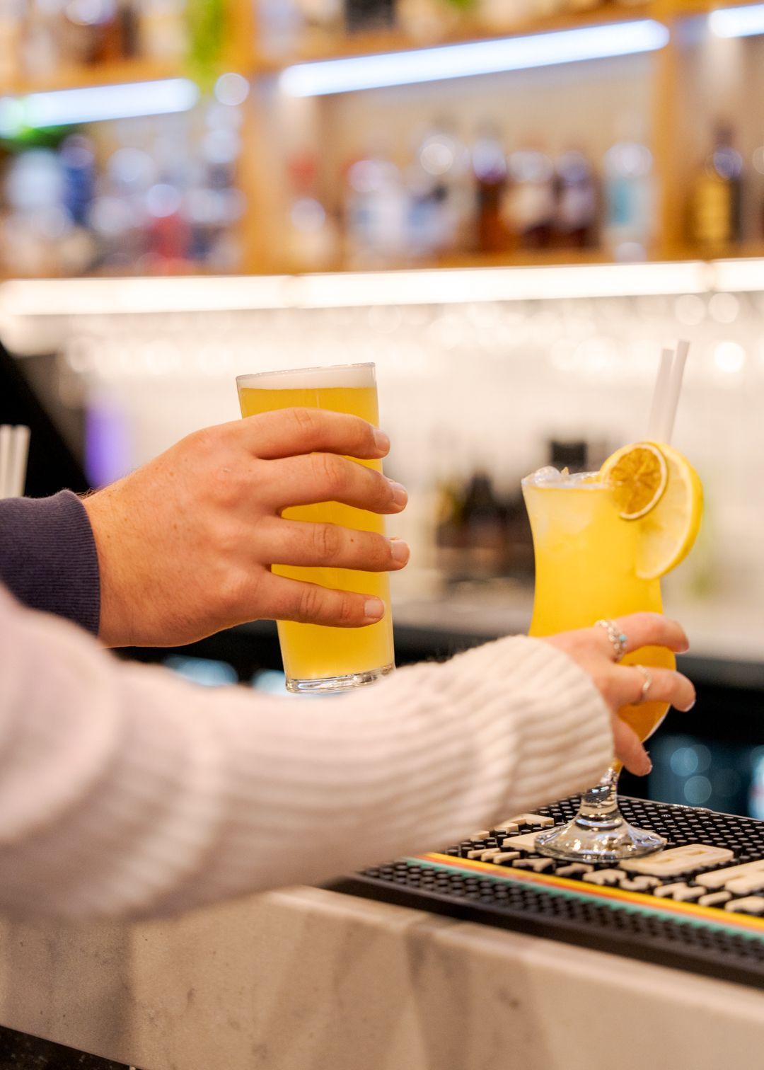Two people clinking orange cocktails at a bar counter, one garnished with a lemon slice.