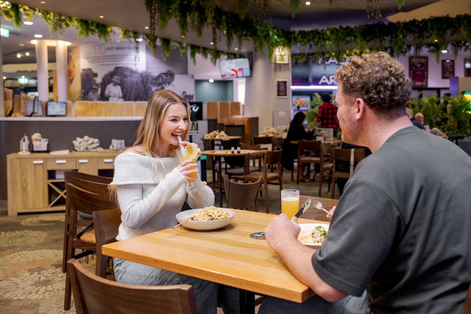 Two people chatting at a wooden table in a cosy café, with drinks and warm hanging lights.
