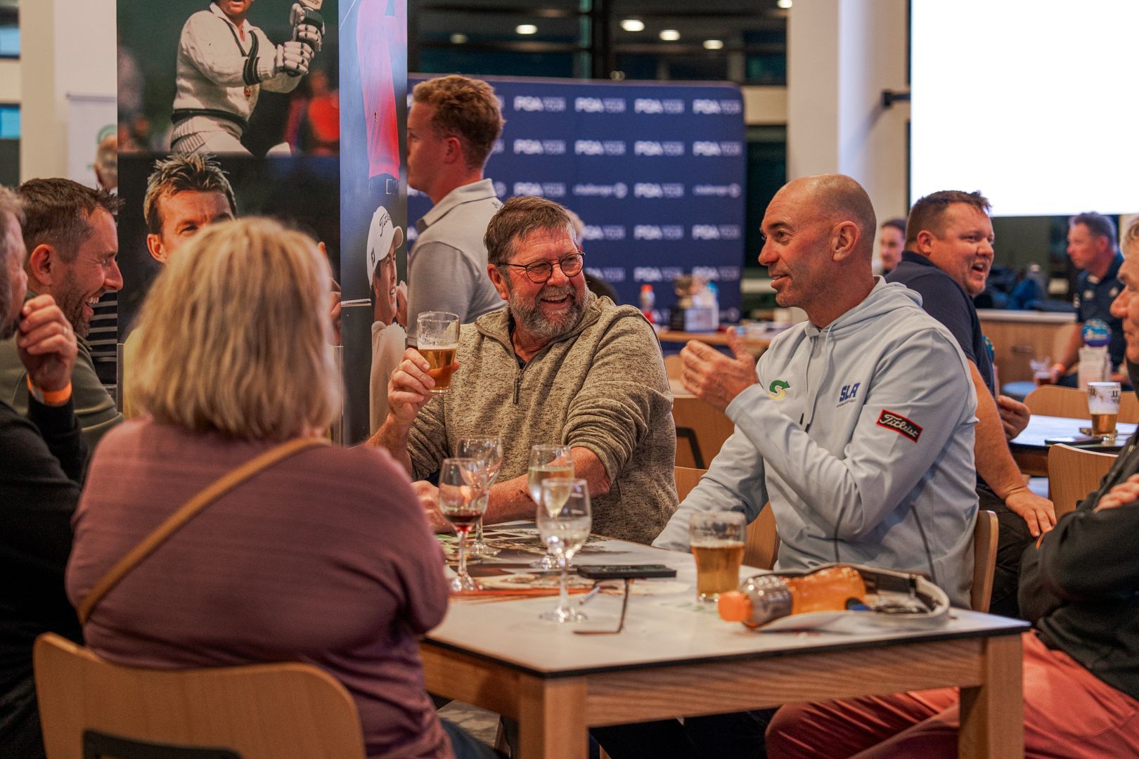 People chatting at a table in a busy indoor social event, with drinks and a blue branded backdrop.