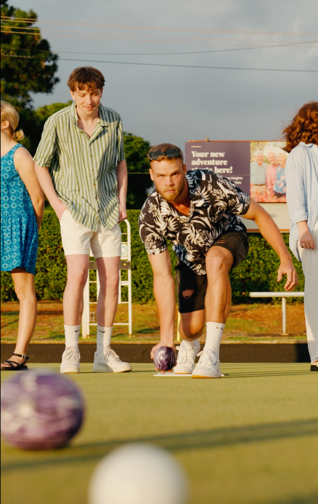 Two people crouch on a lawn bowling green, focusing on a purple ball in the foreground.