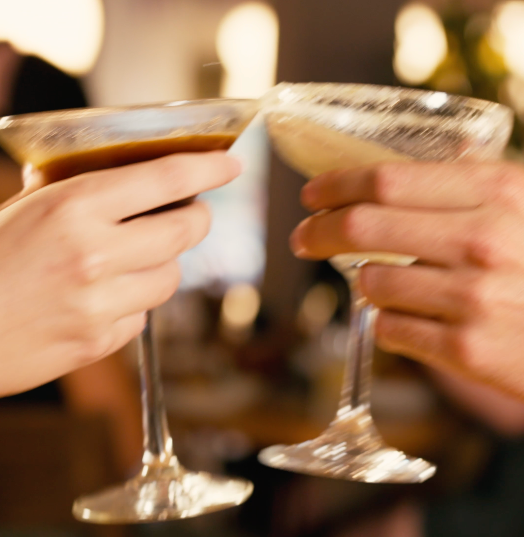 Two hands clink champagne flutes in a warm, blurred indoor setting.