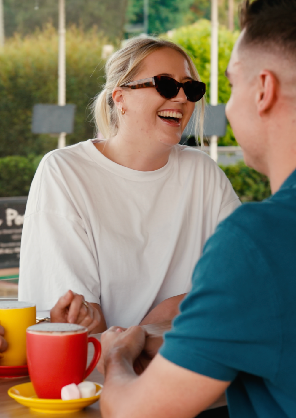 Two people laughing at an outdoor café, with colourful cups on the table.