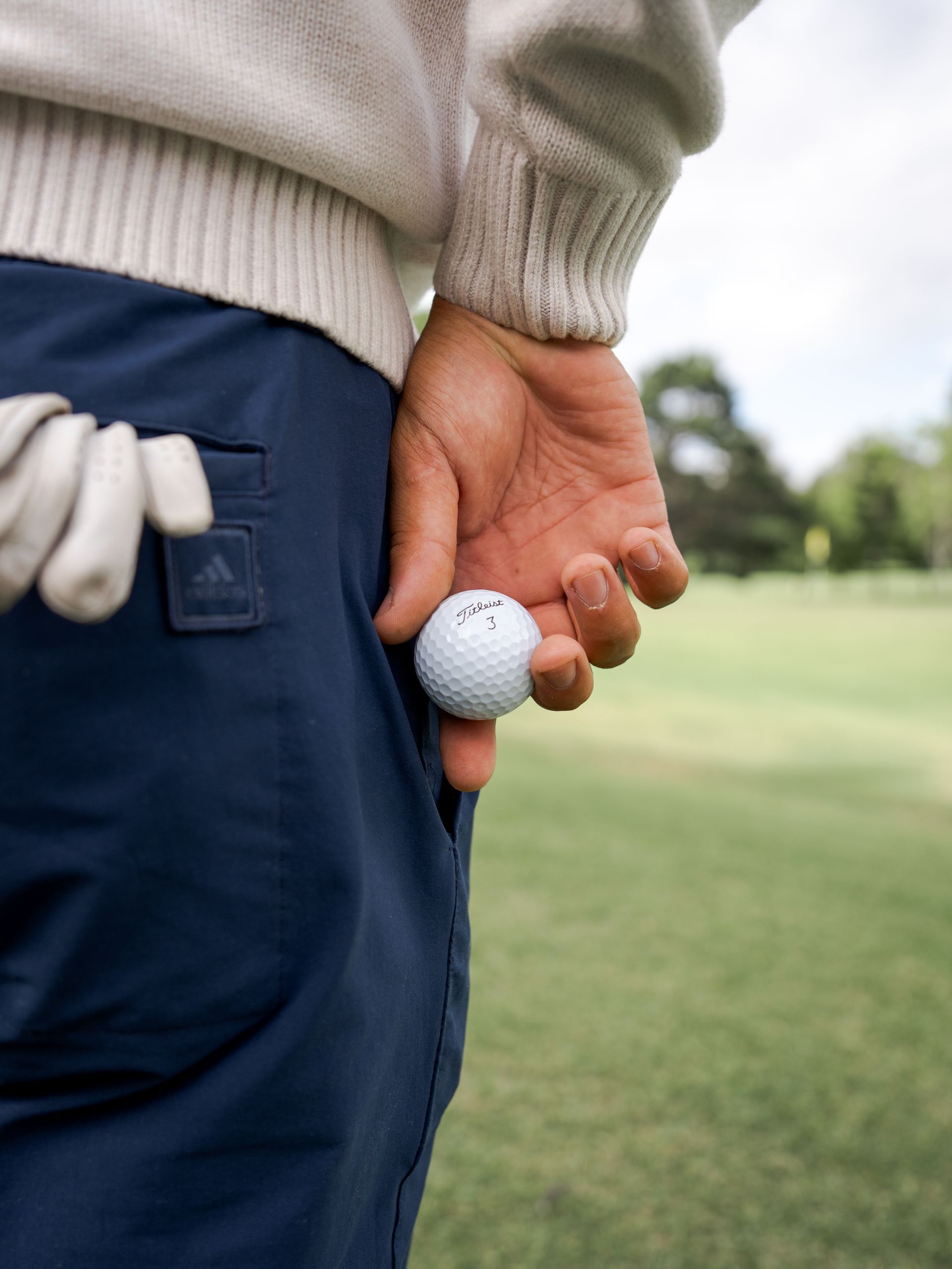 Golfer holding a golf ball beside a green fairway, with tees in a back pocket
