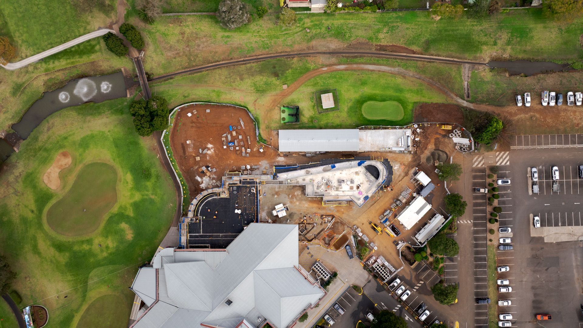 Aerial view of a construction site beside a golf course and parking lot, with buildings and machinery.