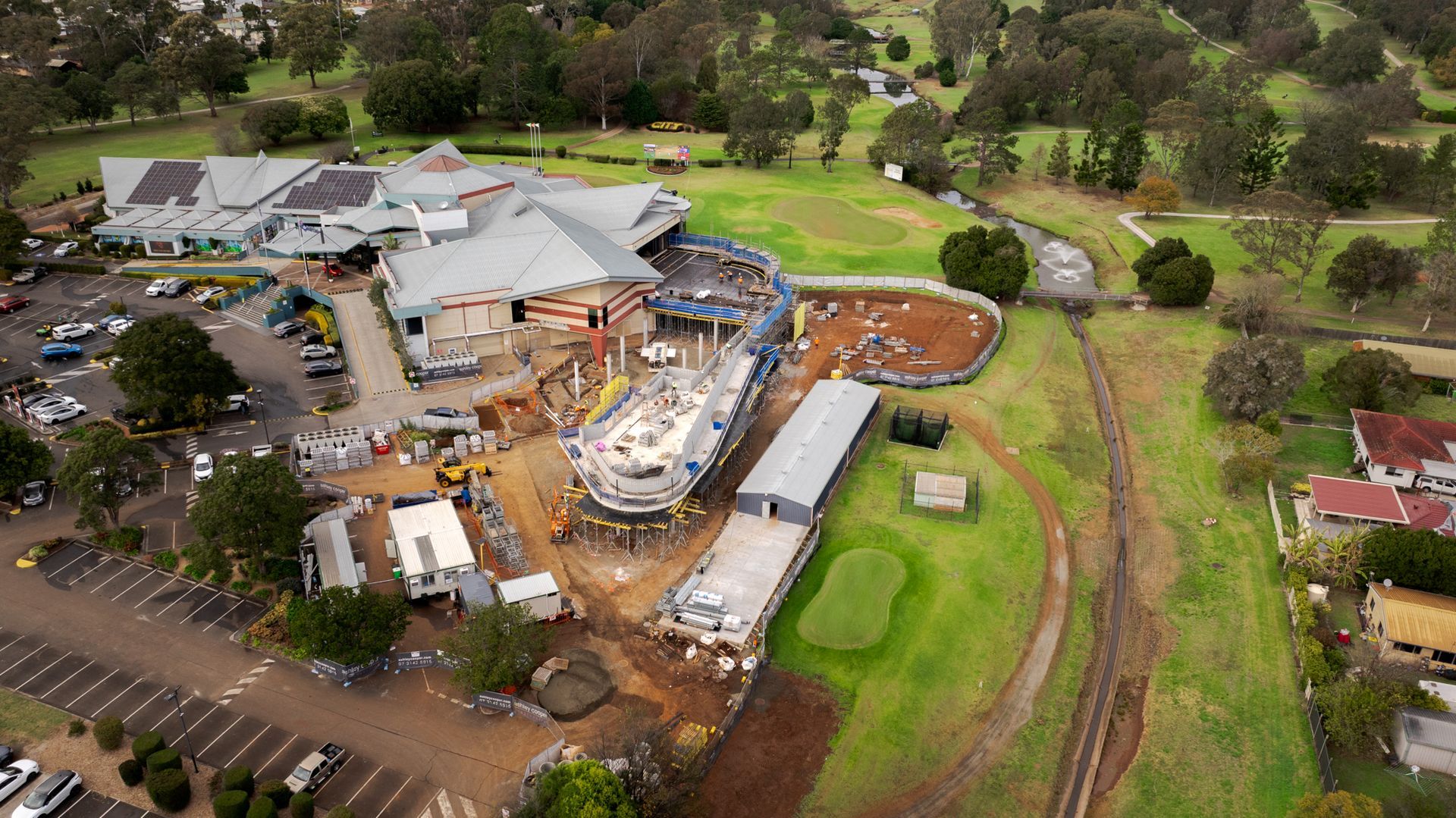 Aerial view of a construction site beside green golf courses, with buildings and earthworks.