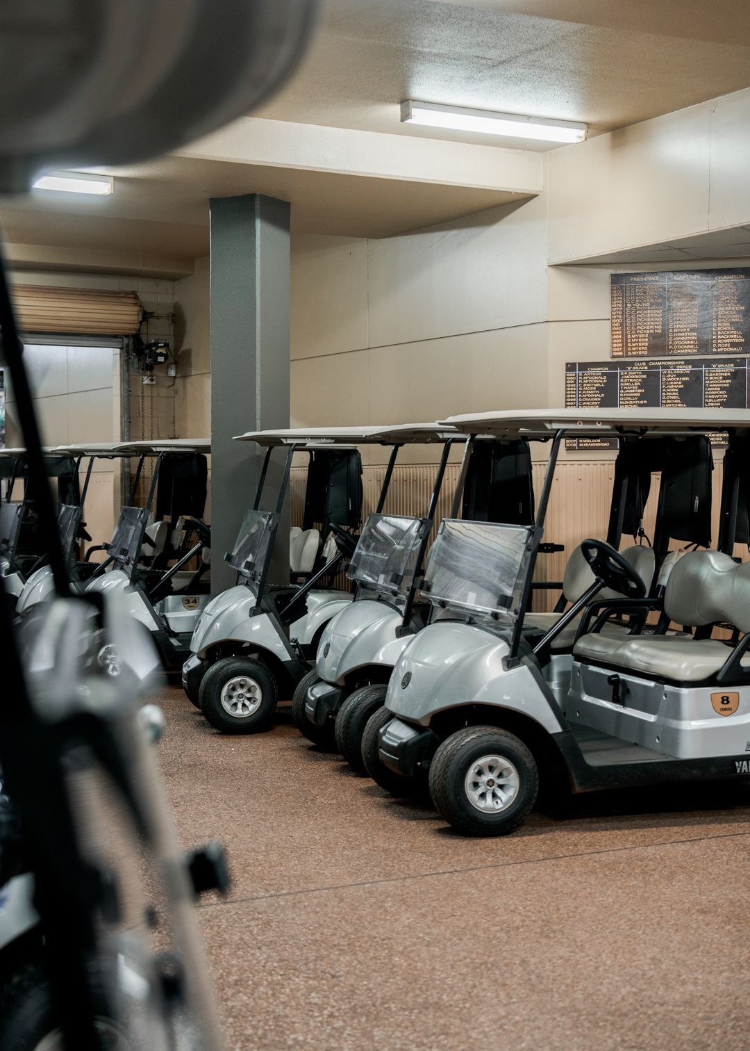 Golf carts parked indoors in a storage area under fluorescent lights.