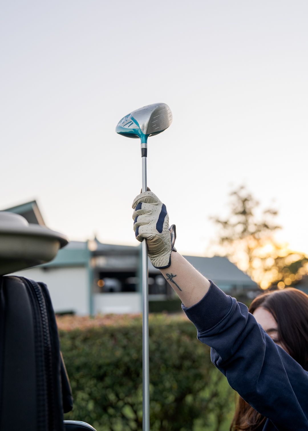 Golfer holding up a club with a blue-and-silver head against an outdoor sunset sky