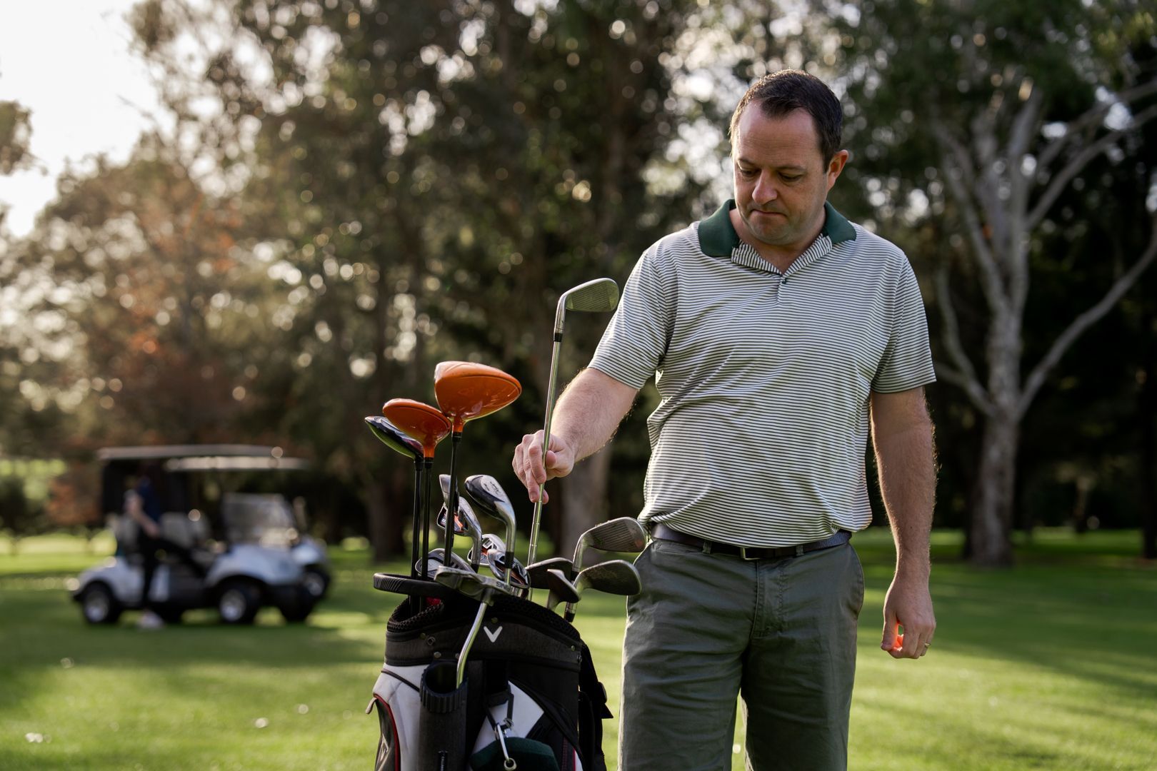 Golfer standing beside a golf bag on a green course, with a golf cart in the background.
