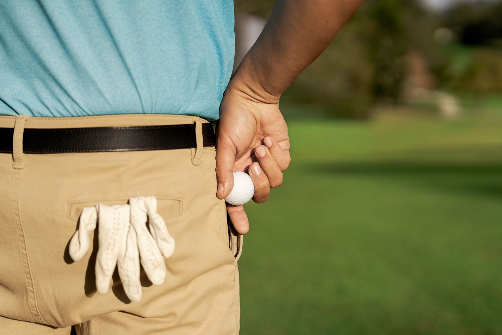 Golfer in blue shirt and khaki pants holding a golf ball and glove on a green course