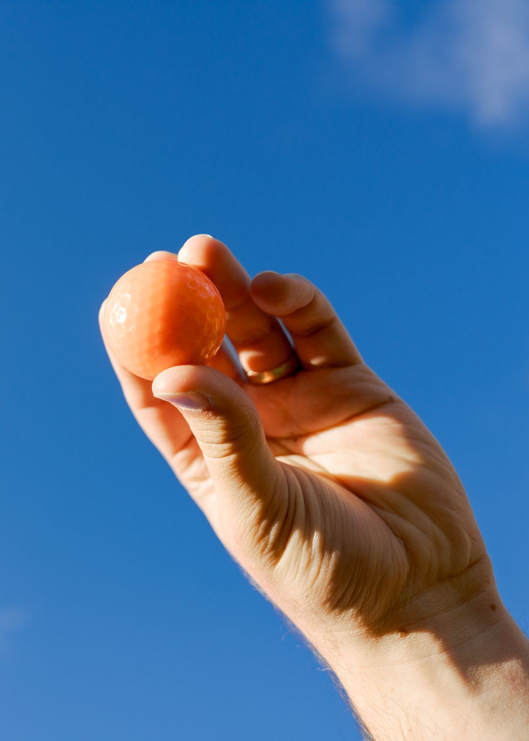 A hand holding a small orange golf ball against a bright blue sky.
