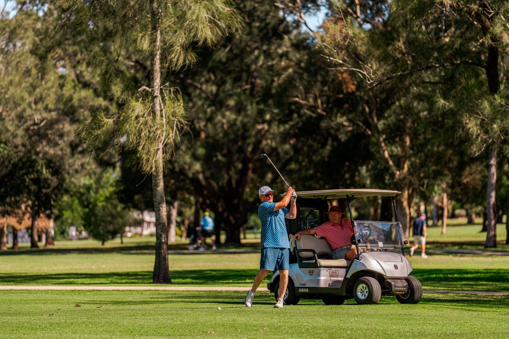 Golfer swinging beside a golf cart in a park-like golf course with trees and green grass