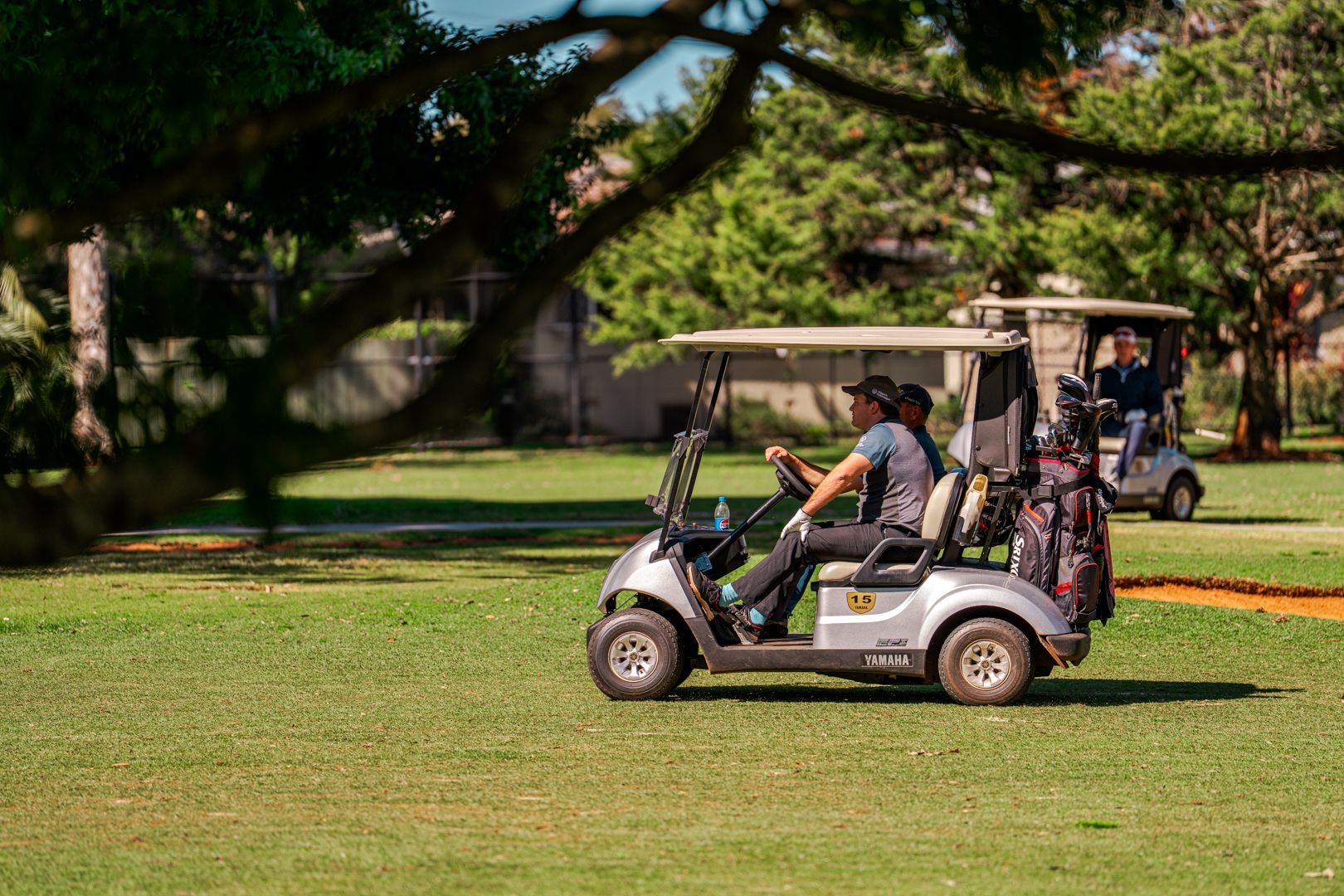 Golf cart with passengers driving across a grassy course under trees
