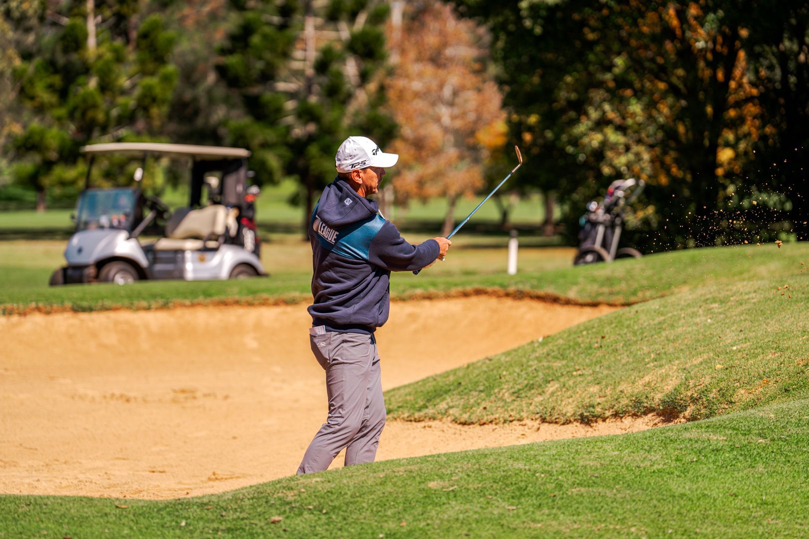 Golfer standing on a course, holding a club near a sand bunker, with carts in the background