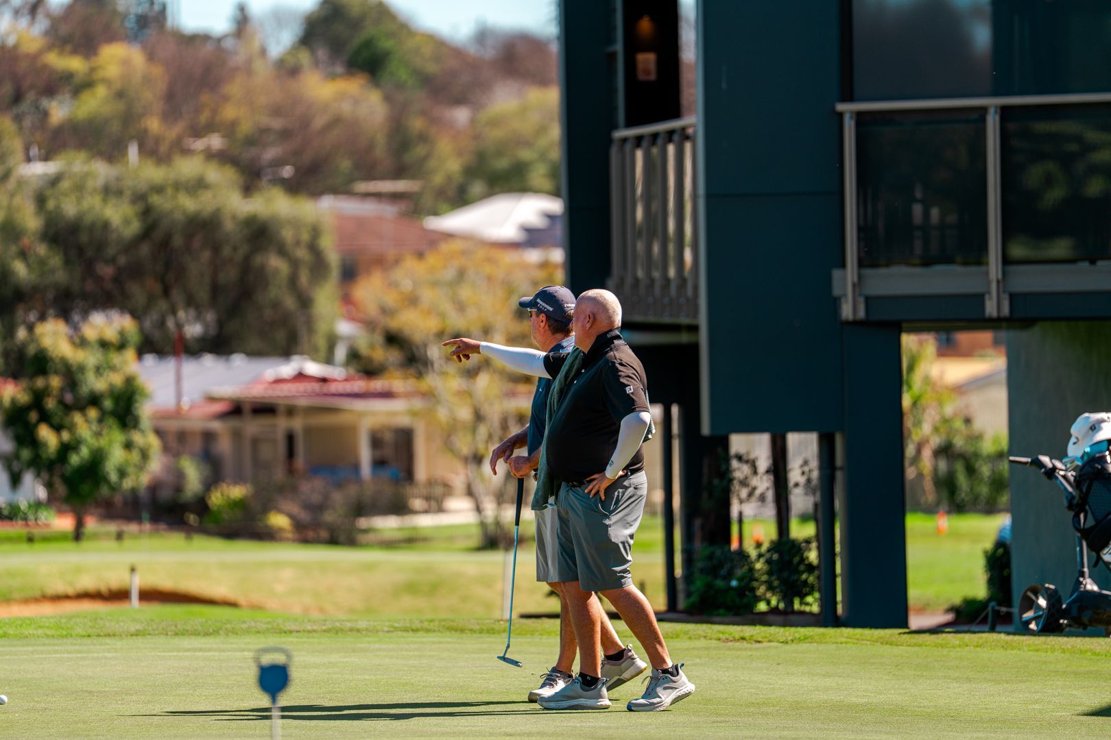 Two golfers standing on a green, one pointing toward the course near a clubhouse.