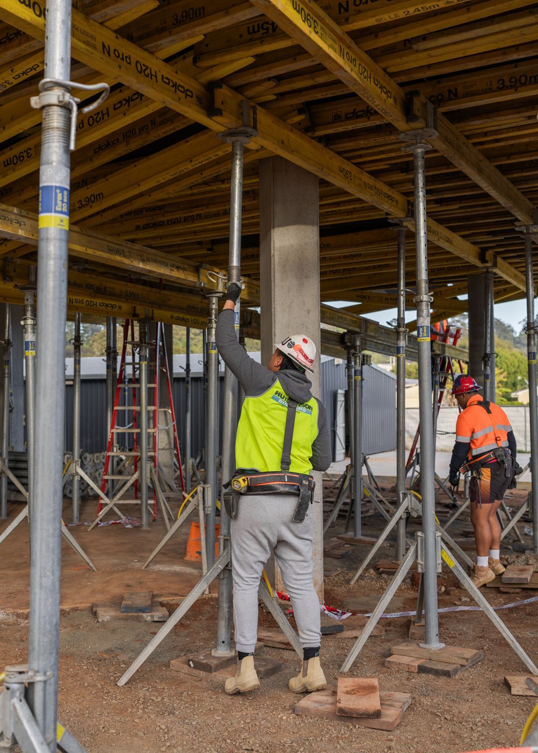 Construction workers under a scaffolded concrete slab, one in a yellow vest and hard hat adjusting a support pole.