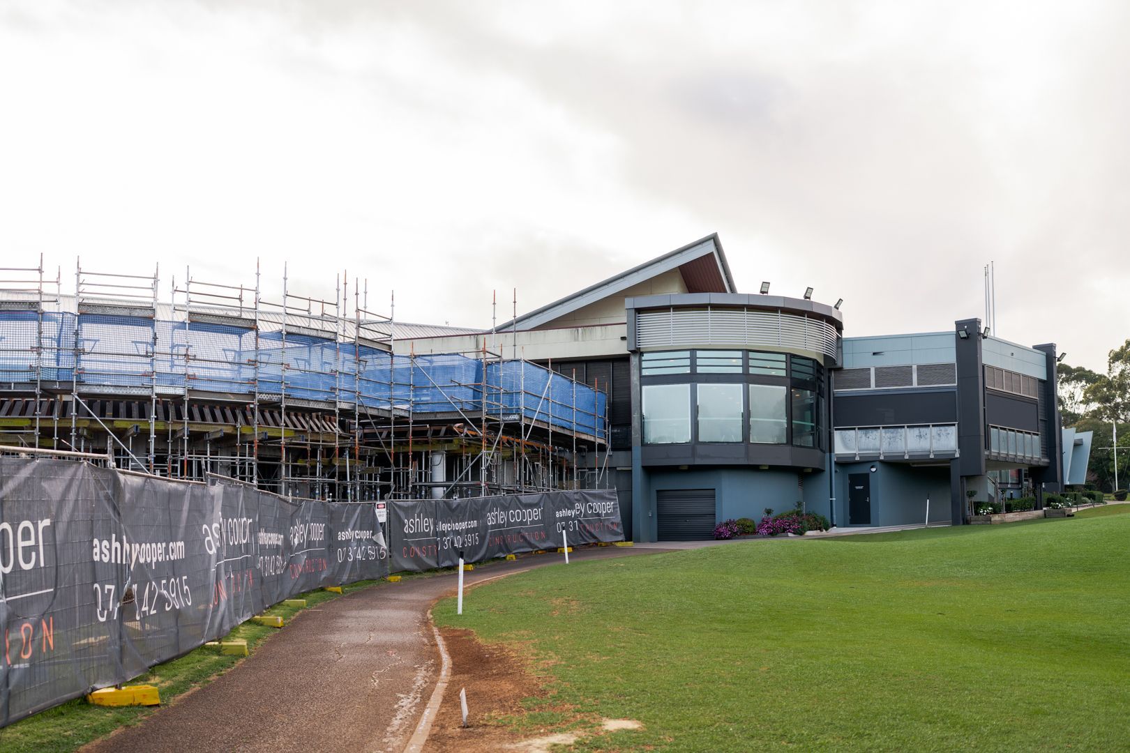 Modern building with scaffolding beside a grassy field and dirt path under cloudy sky