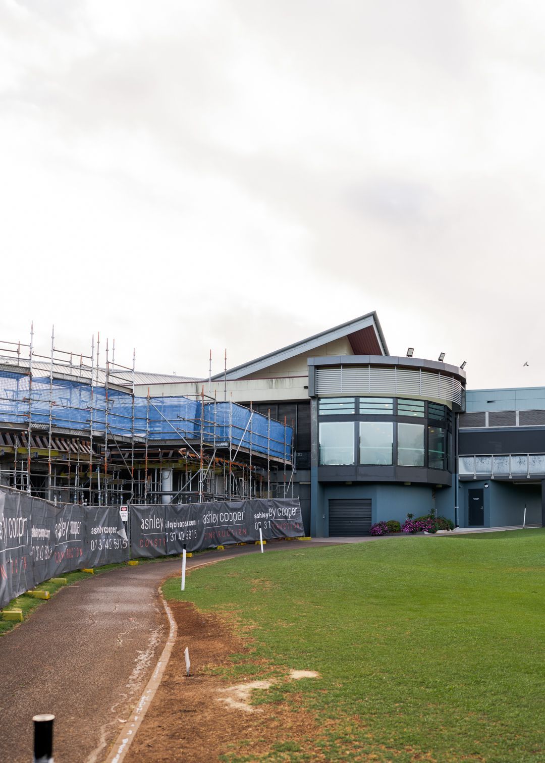 Pavilion beside a grass field, with scaffolding and blue tarps on the left under a cloudy sky