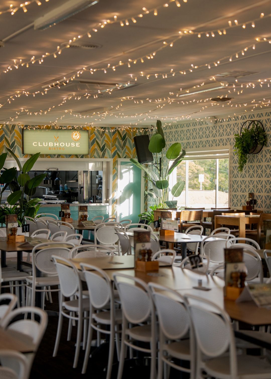 Bright, plant-filled café with white chairs, string lights, and patterned walls