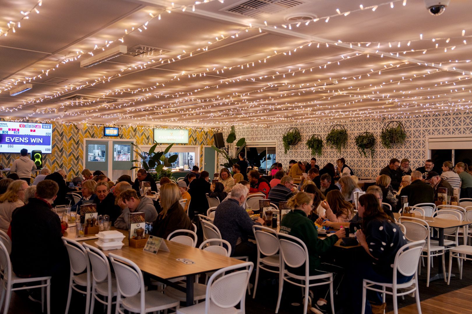 Crowded indoor event with people seated at tables under warm string lights in a decorated hall