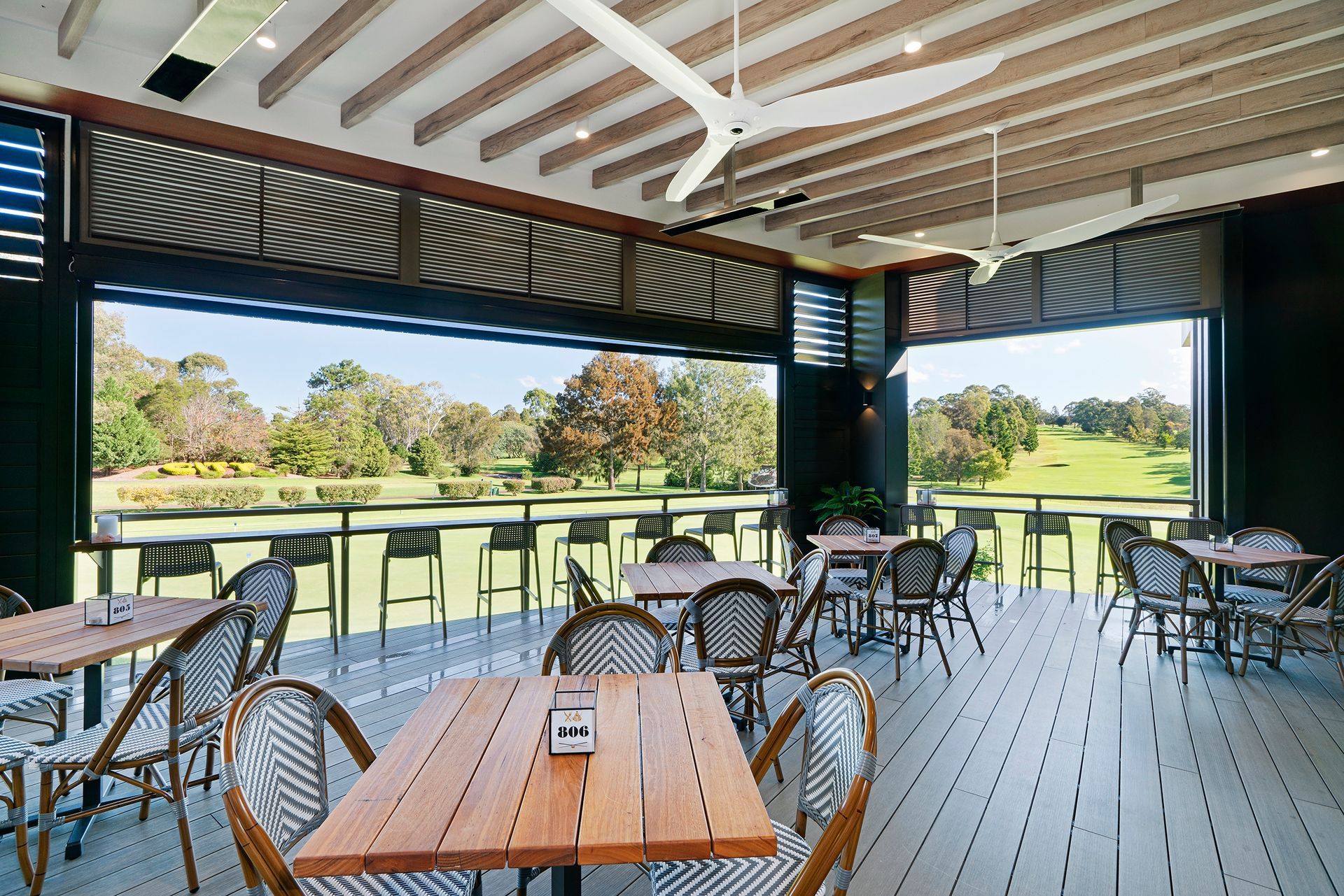 Open-air café patio with wooden tables, metal chairs, and a view of the green golf course.