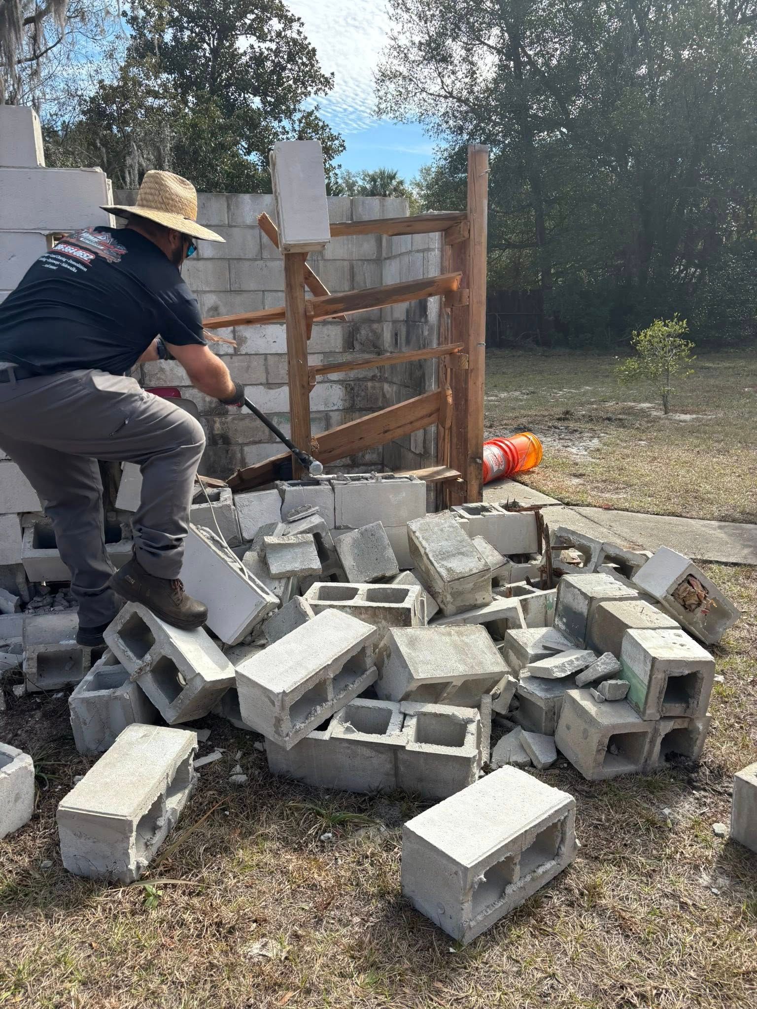 A person wearing a straw hat and dark work clothes uses a pry bar to remove concrete blocks from a wall.