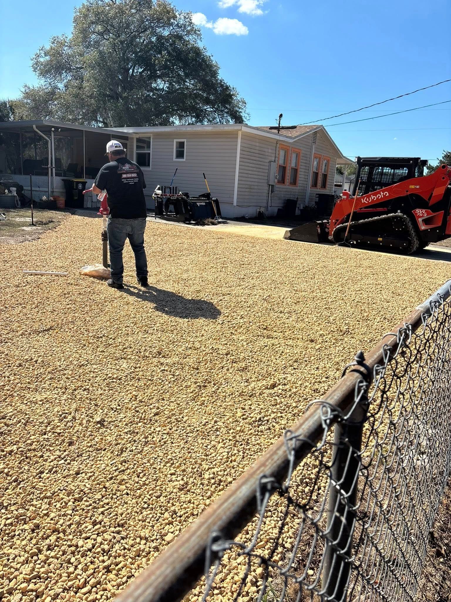 A worker uses a plate compactor on a newly graveled yard in front of a house, with a skid steer loader nearby.