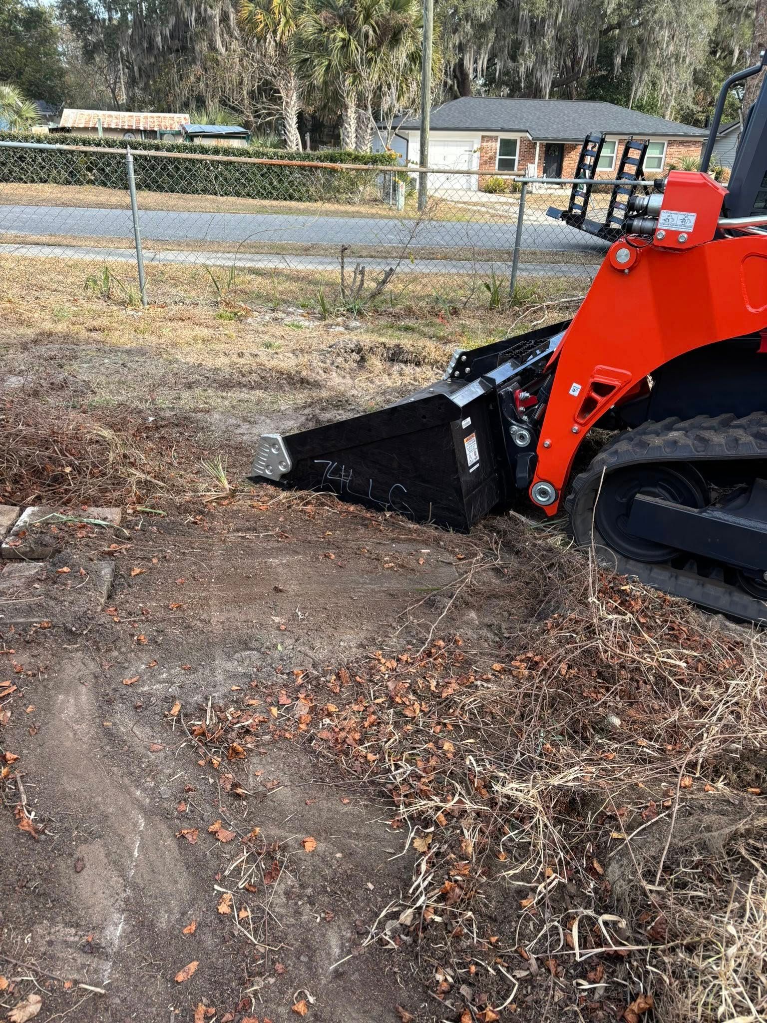 A red skid steer loader with a black bucket sits on a dirt lot, clearing leaves and debris near a residential fence.