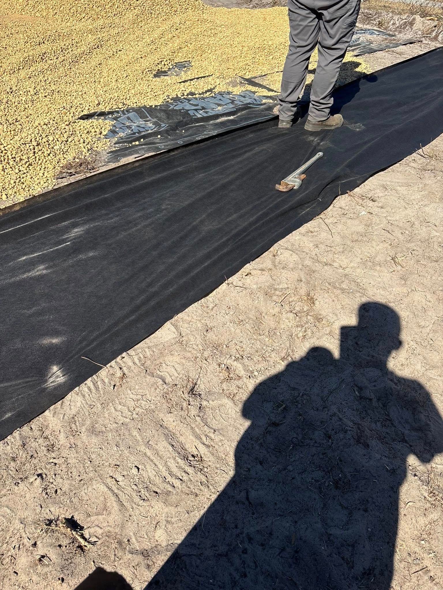 A person stands on a black landscape fabric strip next to a pile of light-colored gravel on a dirt ground in sunlight.