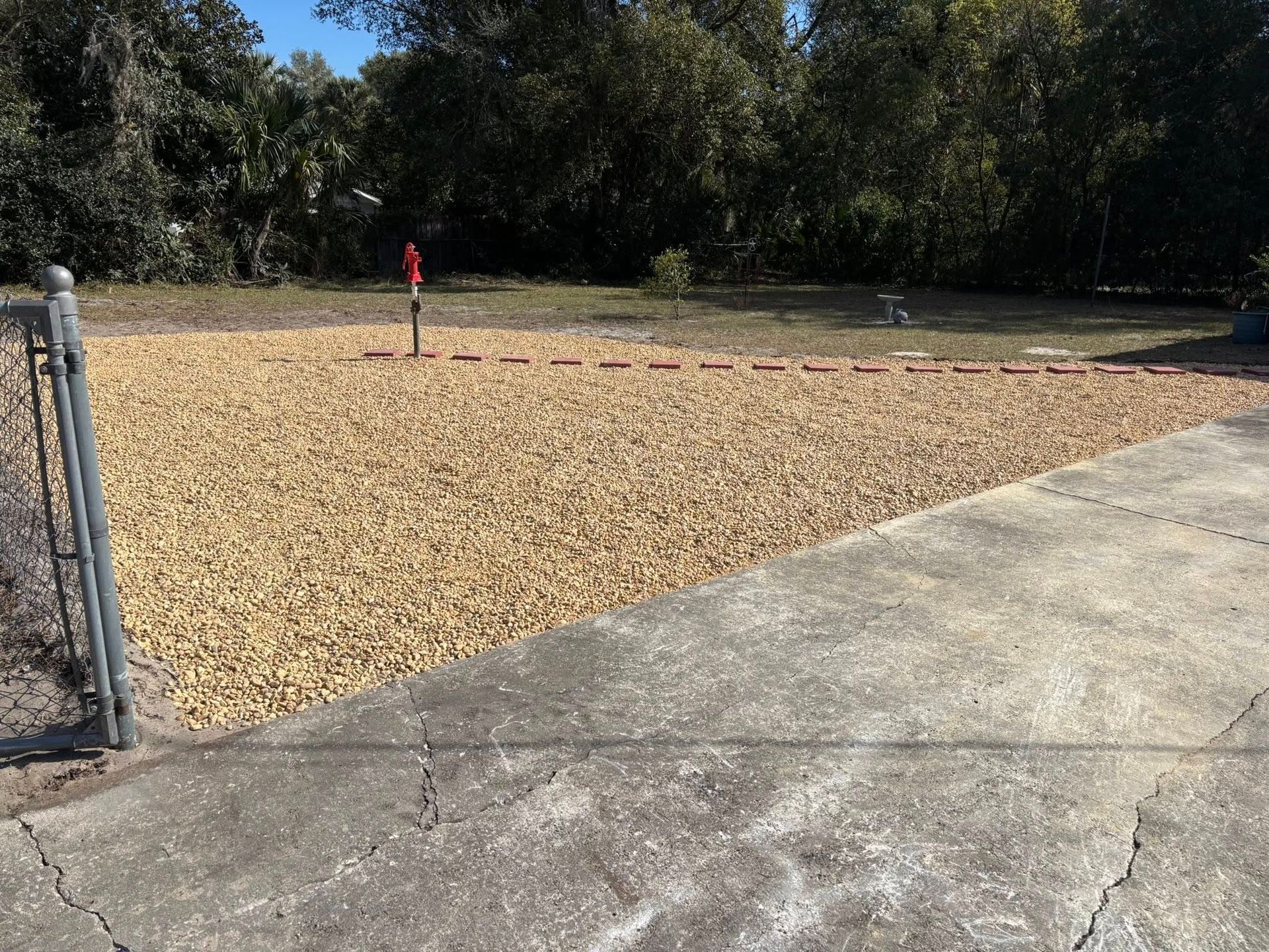 A gravel yard borders a concrete driveway, separated by a red brick edging line, with a metal fence on the left.
