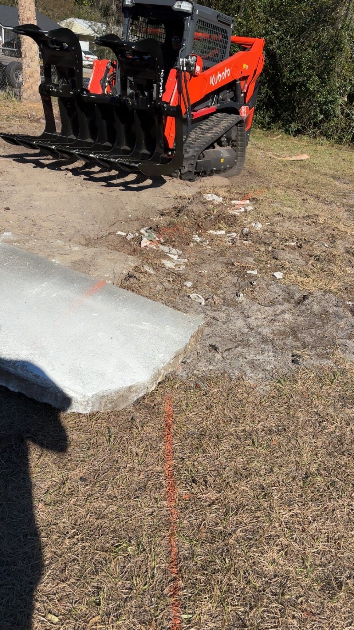 An orange skid steer loader with a grapple attachment parked on a dirt lot next to a concrete slab.