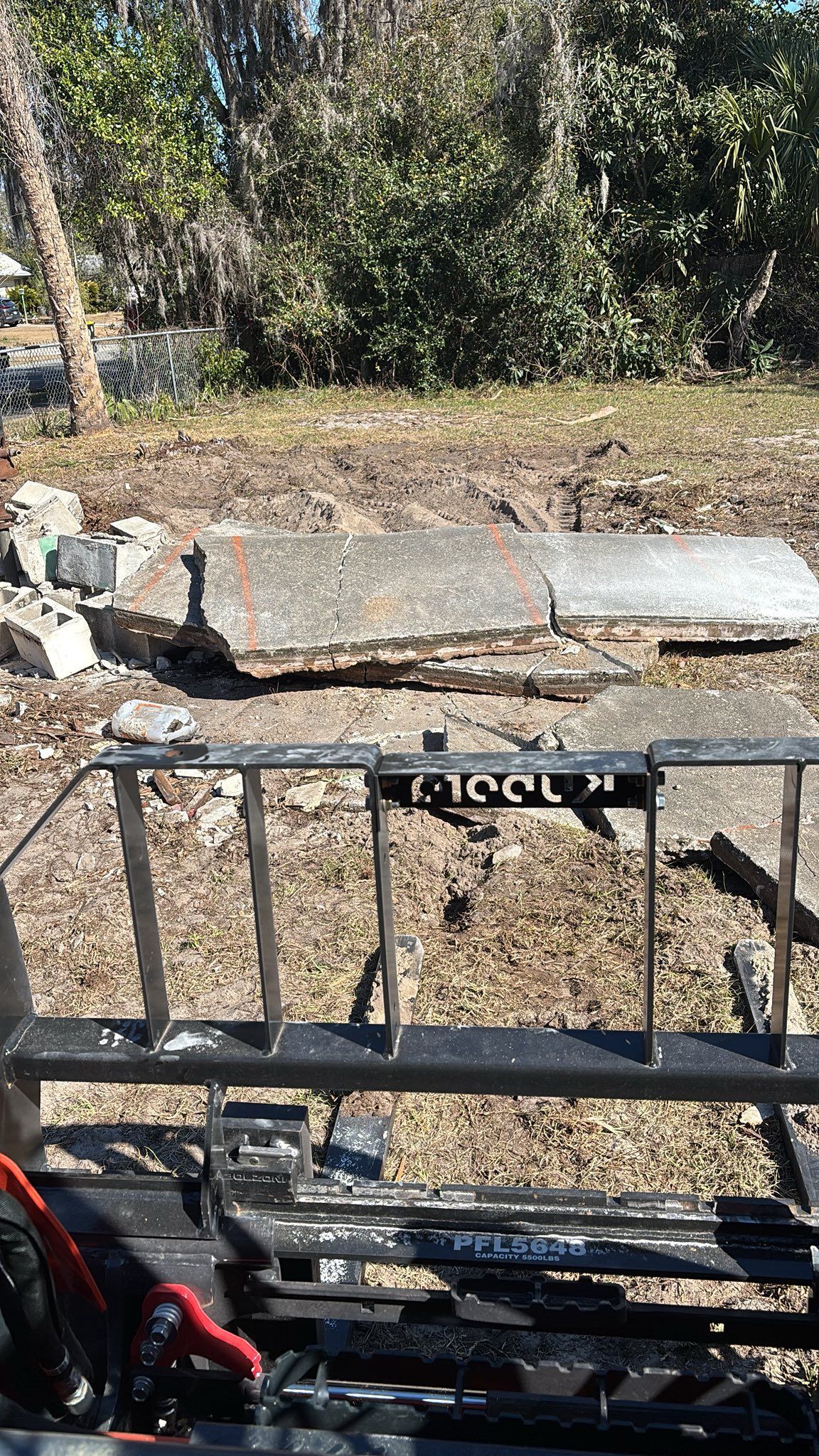 A first-person view from a tractor looking out at a pile of broken concrete slabs on a dirt lot near some trees.