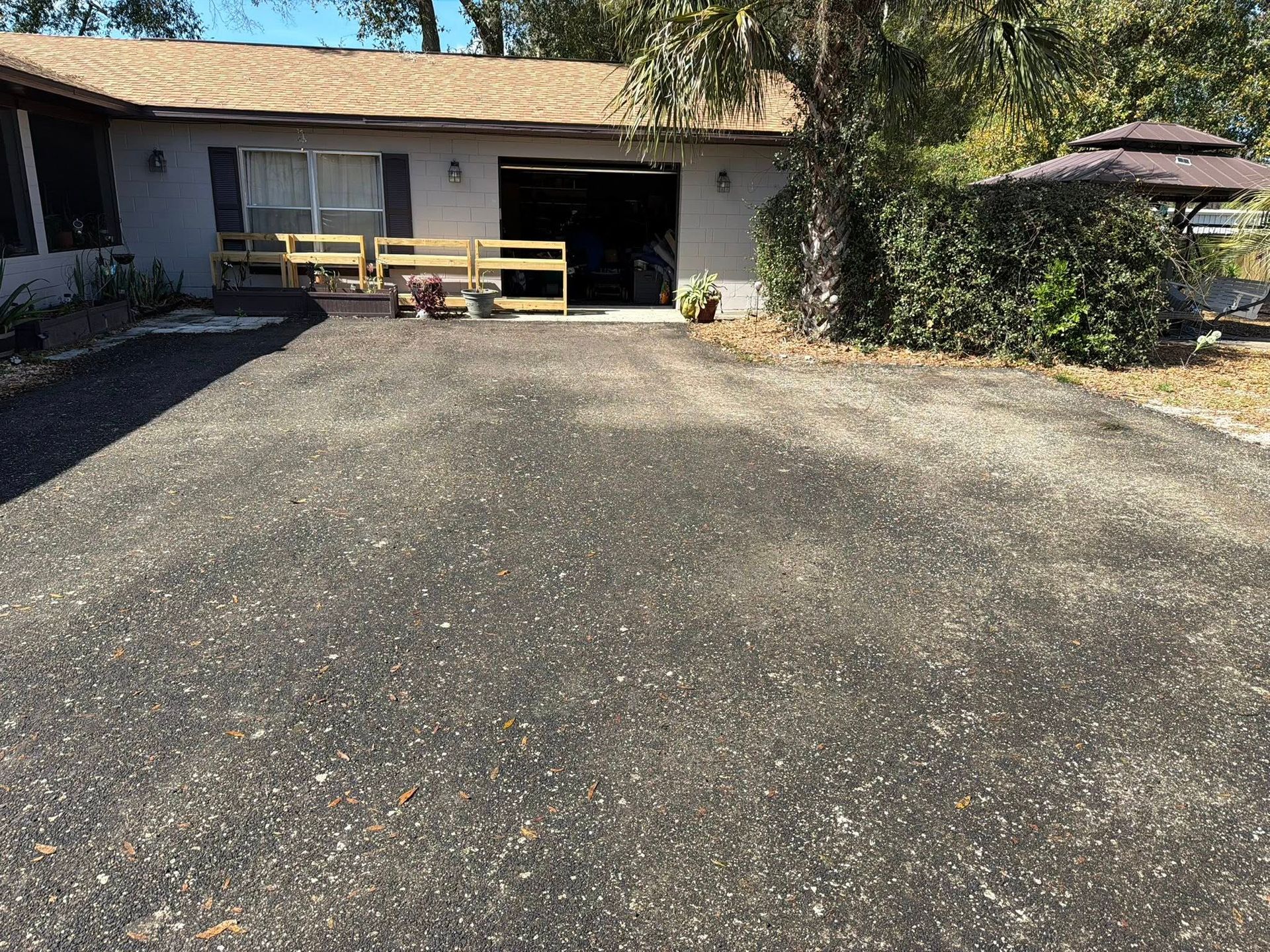 A gravel driveway leads to a single-story home with a garage, patio furniture, and a gazebo on a sunny day.